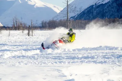 A high-action shot of a snowmobiler carving through deep powder, kicking up a large cloud of snow.