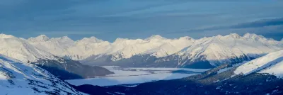 A massive panoramic view looking down into a broad, snow-filled valley nestled between towering mountain ranges in Alaska.