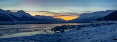 A stunning sunset over a frozen, ice-covered river, with snow-covered mountains in the background and a vibrant orange glow on the horizon.