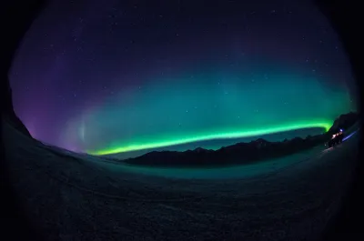 A fish-eye panoramic view showing a vibrant green aurora band arching across the sky from horizon to horizon over a snow-dusted landscape.
