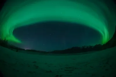 A circular fish-eye view with a wide green aurora arc filling the frame over a vast snowy plain.