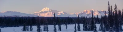 A wide panoramic view of a snow-covered mountain range at sunset, with the peak of Denali glowing in the soft pink light.