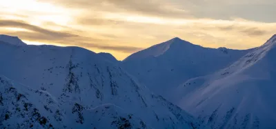 A high-altitude shot of snow-covered mountain peaks in Alaska under a soft golden sunset sky.