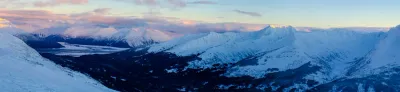 A vast panoramic view of a snow-filled valley and mountains in Alaska at sunset, with soft pink and purple clouds in the sky.