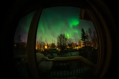 A view through a fish-eye lens from a balcony looking out at a vibrant green aurora over a snowy residential area.