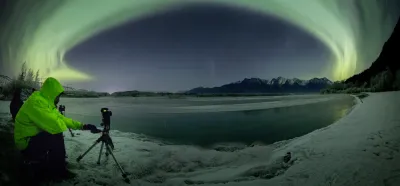 A person in a bright green jacket setting up a camera on a tripod to photograph a massive green aurora arc over a snowy riverbank.
