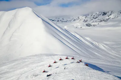 A high-altitude shot looking down on snowmobilers on a ridge surrounded by pristine snowfields.
