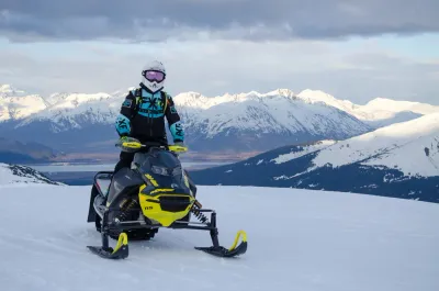 A snowmobiler in gear sitting on their machine, looking out over a vast Alaskan valley.