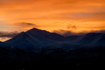 A moody sunset over silhouetted Alaskan mountain peaks, with a bright orange glow in the gaps between the mountains.