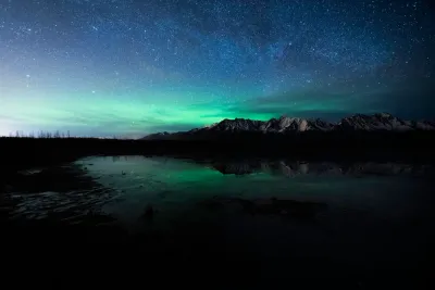 A beautiful night shot of the Aurora Borealis dancing over a snow-covered mountain range, reflected in the Knik River.