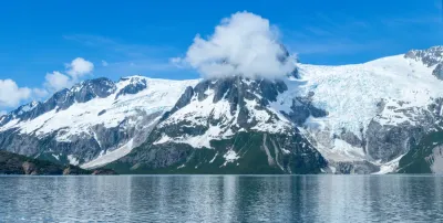 A wide panoramic view of glaciers and snow-covered mountains reflecting in the calm waters of Resurrection Bay near Seward, Alaska.