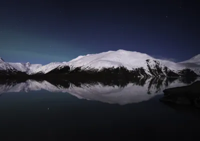 A serene image of a massive snow-covered mountain reflected in a perfectly still, deep blue lake under a starry night sky.