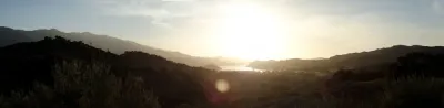 A panoramic view of the Santa Ynez Valley at sunset, with rolling hills silhouetted against a golden sky and a distant lake reflecting the sunlight.
