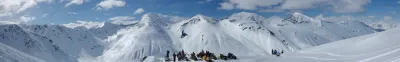 A stunning wide panoramic view of snow-covered mountain peaks with snowmobilers on a ridge.