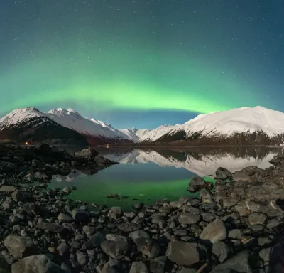 A stunning panorama of a brilliant green aurora band arching over a calm lake and snow-covered mountains, with rocky foreground details.
