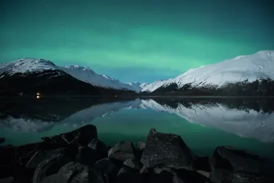 A breathtaking panorama with the green aurora reflecting in an icy lake surface, framed by snowy peaks.