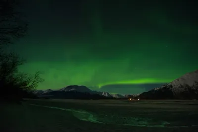 A low-angle shot of the green aurora glow low over a dark mountain range and a wide, frozen river plain.