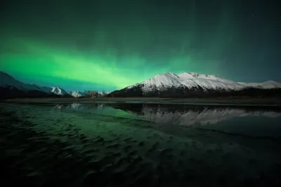 A wide field of green aurora light hanging above a dark mountain range and a vast, flat snowy foreground.
