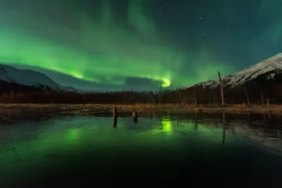 A brighter and more defined green aurora arc over a still lake and snowy peaks, with a beautiful reflection.