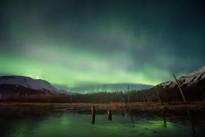 A dramatic wide-angle shot of a green aurora arc stretching across the sky from behind a dark silhouetted mountain range over an icy river.