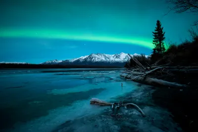 A bright green and pinkish aurora swirl filling the frame over a dark forest and icy riverbank.