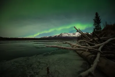 An intense green aurora light arcing over snowy mountains and reflected in an icy river.