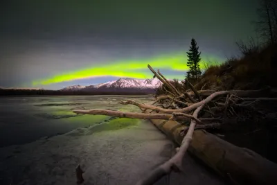 A vivid green aurora arc over a mountain range, with foreground driftwood and ice visible.