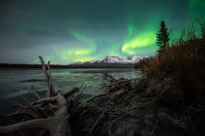 A wide-angle view showing a more diffused green aurora glow over snowy mountain peaks and a foreground river with logs.