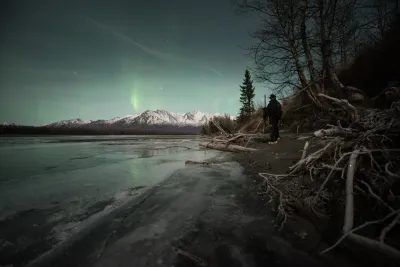 A photographer silhouetted in the corner, overlooking a wide frozen plain and mountains under a vast, soft green aurora sky.