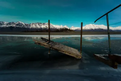 A damaged wooden pier tilted in the ice of a frozen lake, under a deep blue starry sky with a faint green aurora glow and snow-capped mountains.